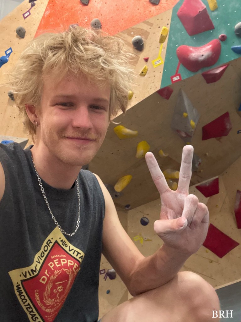 The writer, Brady Hawkins, sitting in front of a climbing wall, displaying a peace sign with his hand.